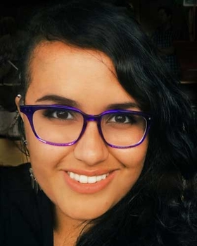 Image of Juliana Pino, smiling afroindigenous person with dark hair, dark eyes, brown skin, purple glasses